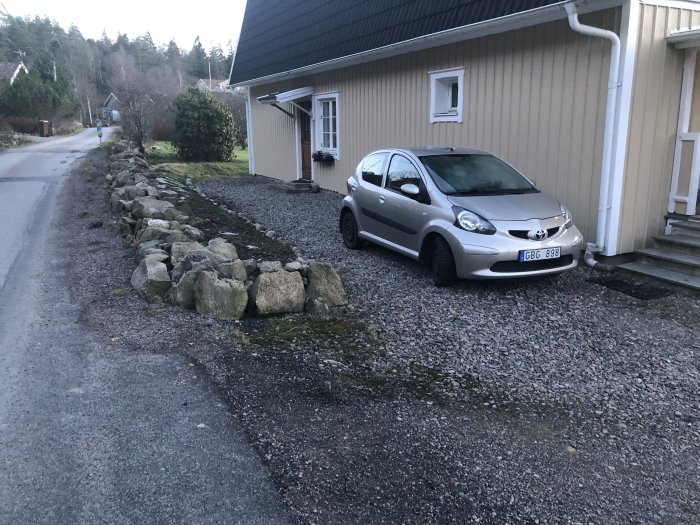 Bild på sidan av ett hus med stengärde, grusdrivväg och en parkerad bil.