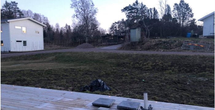 Tomt med sluttning intill gräsmatta, väntande på landskapsplanering, med hus och träd i bakgrunden.