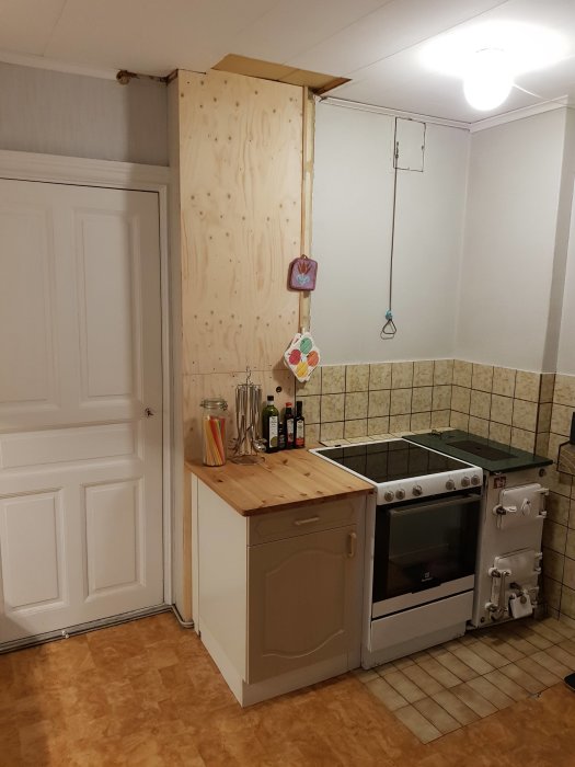 Kök före renovering med upptäckta rör och ledningar bakom en plywoodvägg, intill spis och vit skåpslucka.