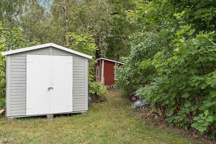 Vit skjul och rött förråd omgivna av gröna träd och buskar på en lummig tomt i Blekinge.