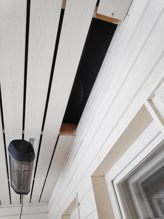 Öppning i en vitmålad trätakstol under en inglasad terass med synlig ventilationsspiral och väggmonterad värmare.