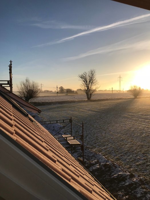 Morgonsol över frosttäckt landskap sett från tak med byggnadsställning och tegelpannor.