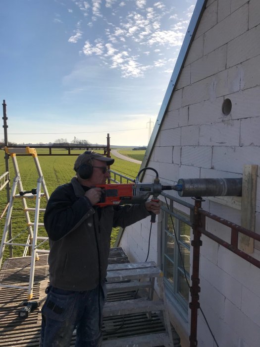 Arbetare installerar ett avloppsrör på en vit tegelvägg, utomhus med landskap i bakgrunden.