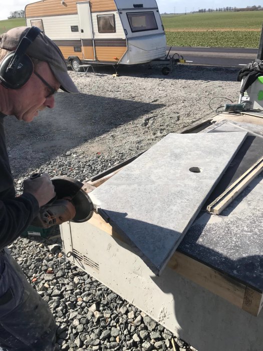 Man som använder vinkelslip på en betongplatta vid byggarbetsplats med husvagn i bakgrunden.