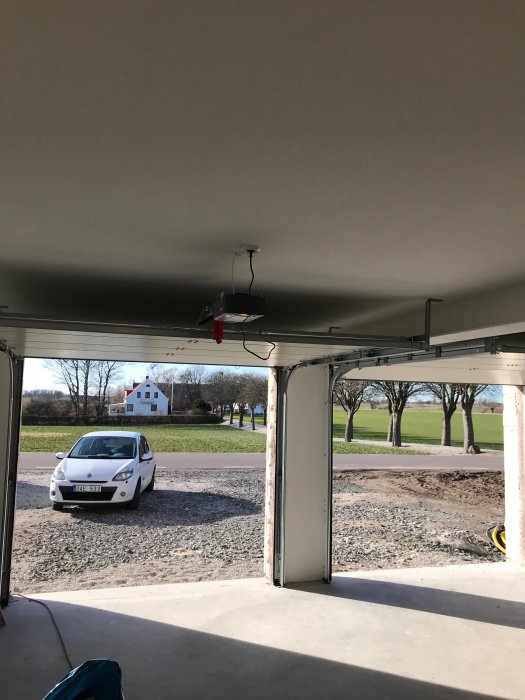 Vy från ett öppet garage mot en vit bil parkerad utanför på en grusplan, med gröna fält och ett hus i bakgrunden.