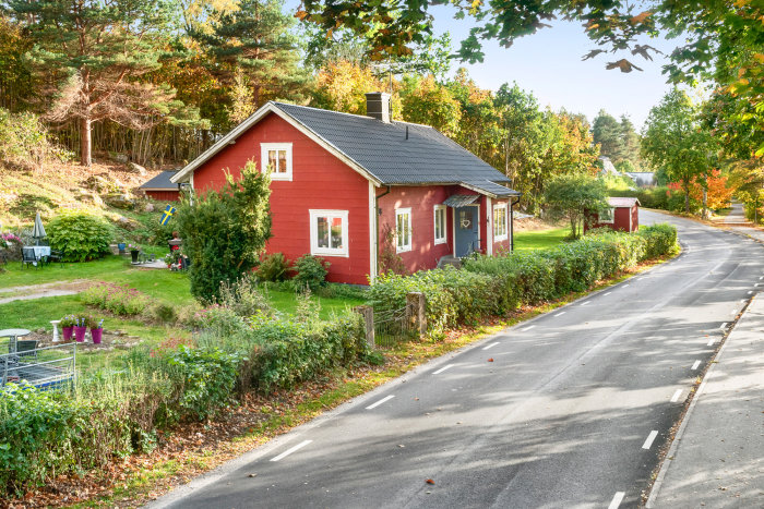 Ett charmigt litet rött hus med svart tak vid en väg omringat av grönska och träd i svenskt landskap.