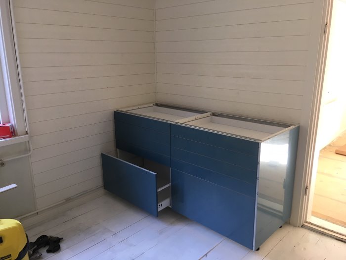 Renoverat kök med vit panel på väggarna och blått Ikea-bänkskåp under montering, ljust målat golv.