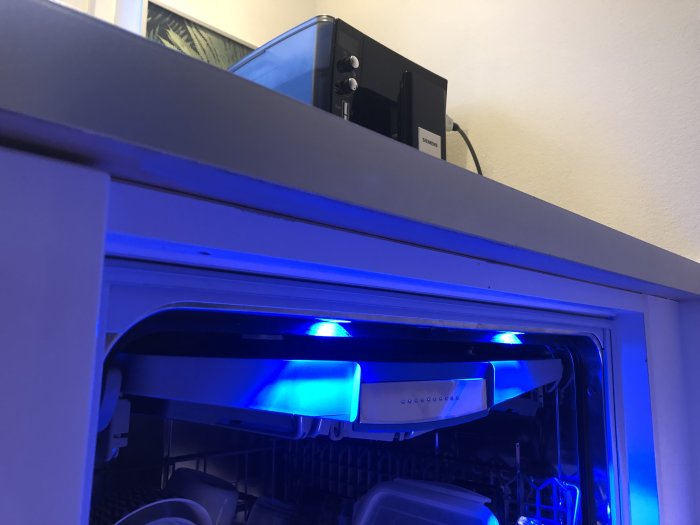 Diskmaskin öppen med blått ljus och skåp ovanför med elektronik, i ett hemmiljö.