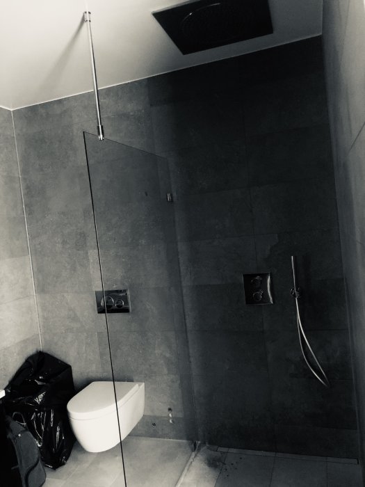 Renoverat badrum med mörkt kakel, vägghängd toalett och dusch med glasvägg.
