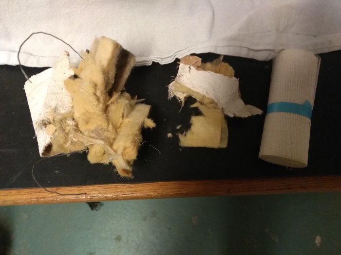 Delar av isoleringsmaterial bredvid en rulle väv på en träyta, möjligt asbestinnehåll under utredning.