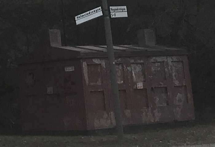 Förvittnad stor röd elstation vid en vägkorsning med gatuskyltar, solljus och träd i bakgrunden.