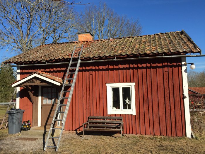 Röd stuga med tegeltak och stege lutad mot fasaden, omgivet av träd under en klarblå himmel.