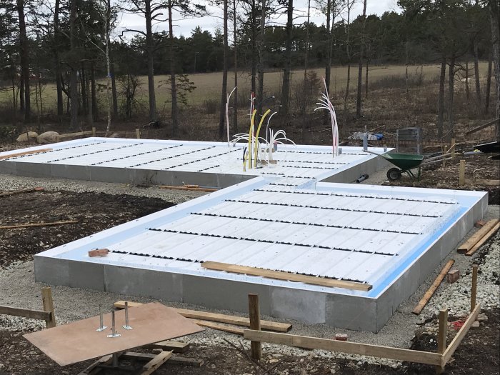 Grundläggning för hus med cellplastisolering och utlagda golvvärmeslangar på en byggarbetsplats i skogsmiljö.