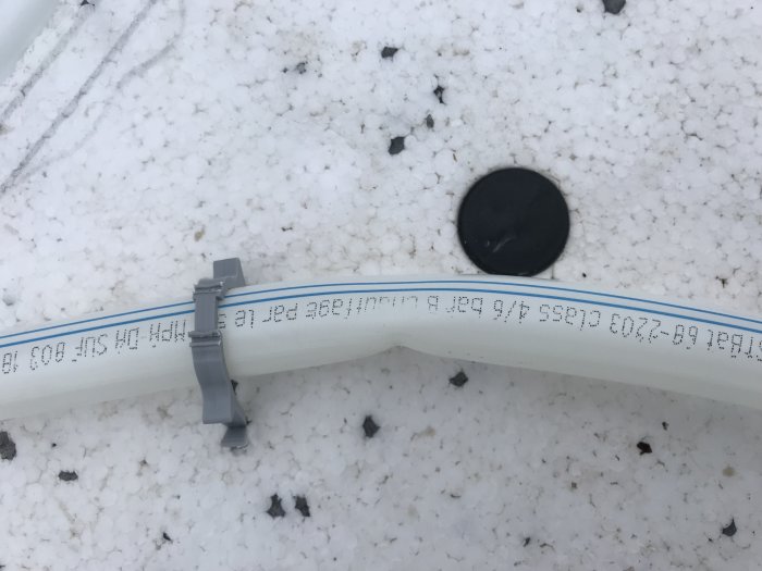 Golvvärmeslang med veck skadad på en yta med småsten, säkrad med kabelklammer på ett underlag av vit cellplast.