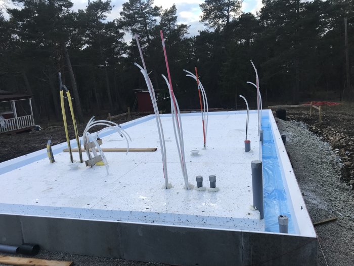 Byggplats med cellplast och utlagda rör för avlopp och golvvärme i förarbetet för en grund.
