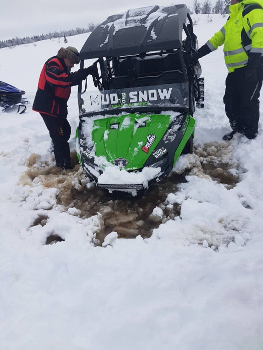 Två personer försöker dra ut en grön och svart snöskoter som fastnat i snön i Lofsdalen.