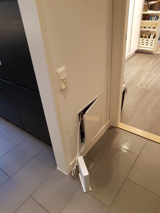 Öppen lucka i väggen visar nätverksutrustning, med en router på golvet och kablar som går in i väggen.