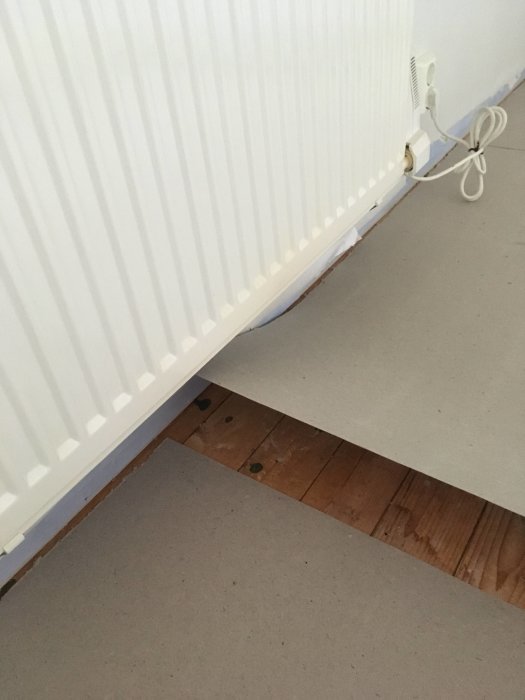 Uppriven del av laminatgolv som visar underliggande plankgolv och grålumppapp nära vägg och radiator.