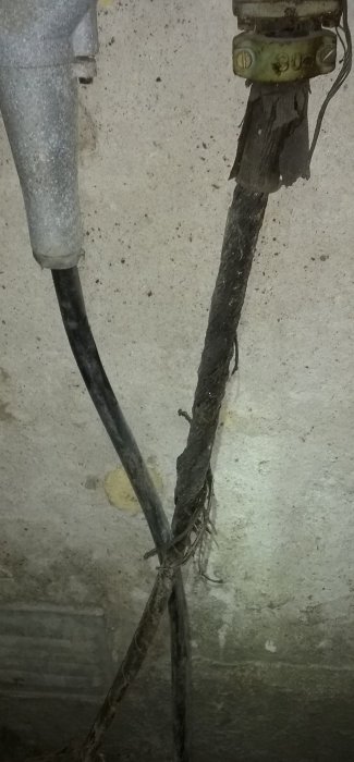 Sliten tyglindad kabel bredvid en plastkabel, fästad i vägg, med tecken på åldring och slitage.