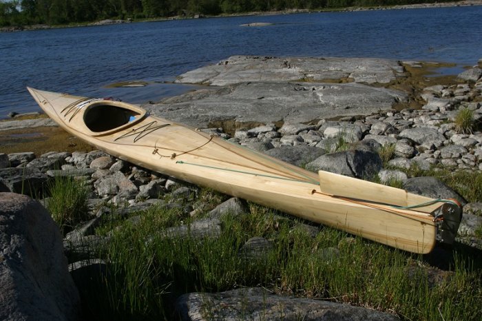 Handbyggd träkajak med epoxibehandling ligger på klippig strand vid vattnet, exemplifierar hantverkskvalitet och hållbarhet.