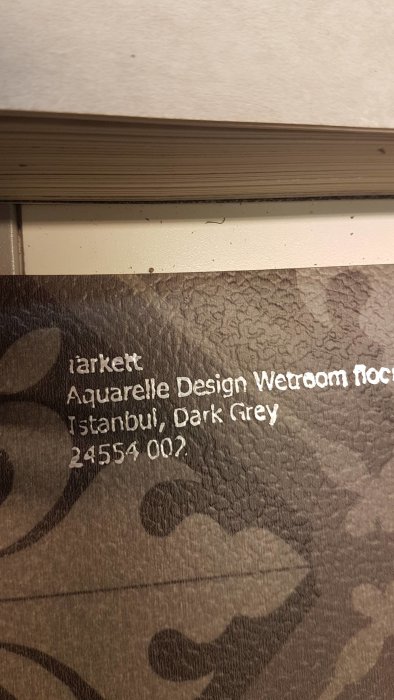 En närbild av en våtrumsmatta med märkningen "Aquarelle Design Wetroom Floor Istanbul, Dark Grey", medan den läggs.