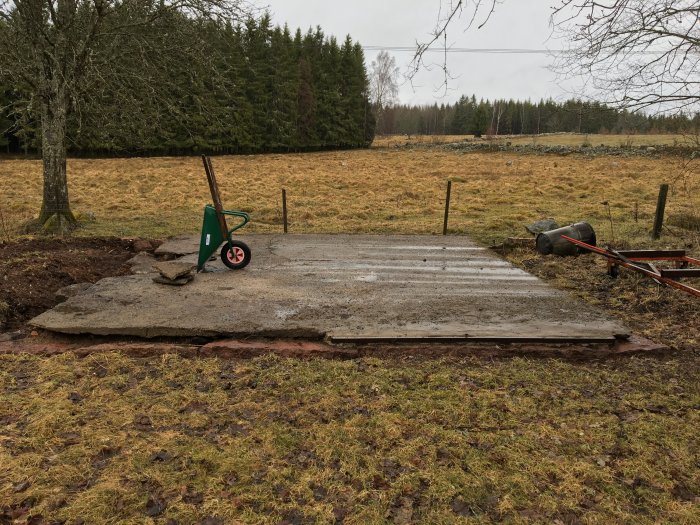 Gammal gjuten betongplatta och grundstenar på en övergiven tomt med skräprydd omgivning.