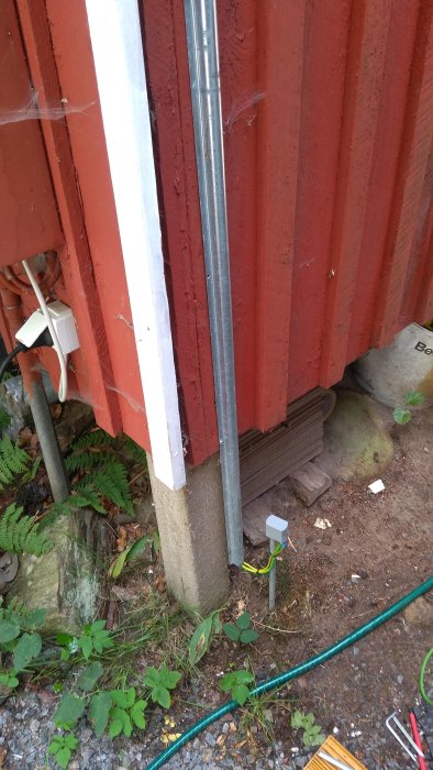 Elinstallation på husvägg med rör och kabel som går till ett uteuttag, omgiven av grönska och ett jordspett.