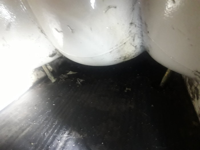 Närbild av golvet under en toalettstol med svart matta och synligt vattenlås.