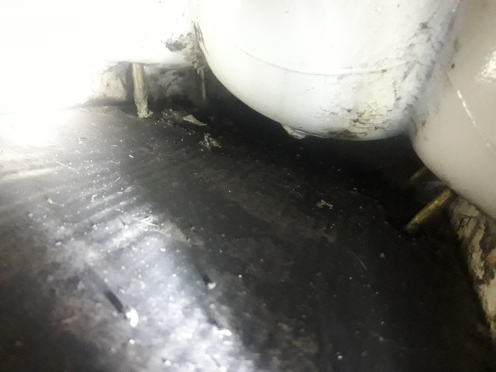 Utrymme under toalettstol med svart matta, synliga rör och vattenlåset.