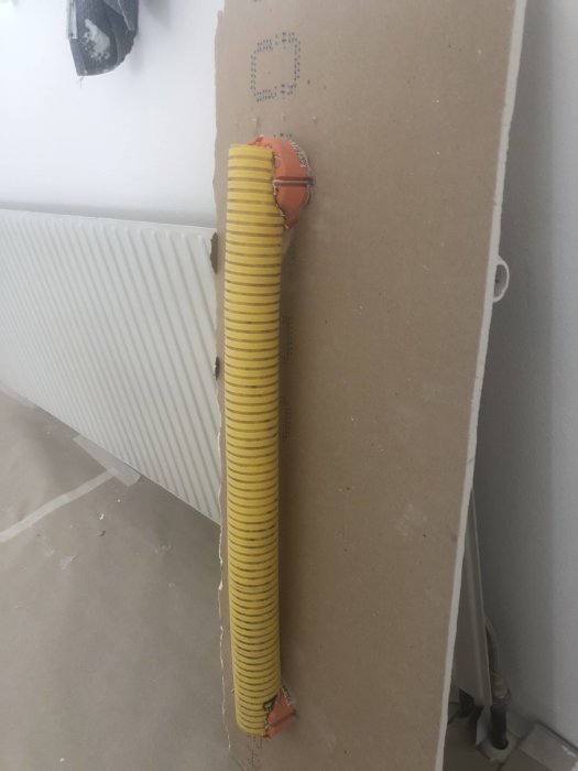 En gul 60mm markslang ledd genom två orange senildosor bakom ett öppnat gipsväggselement nära en radiator.