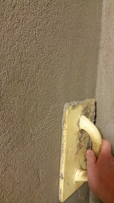 En hand som håller i en gul spackelspade mot en nyspacklad vägg.