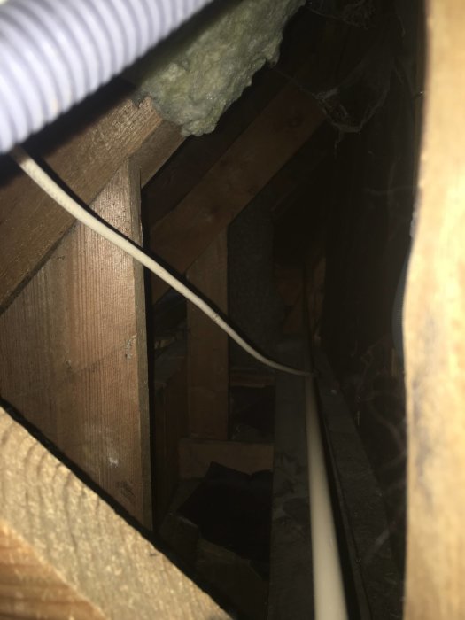 Vy genom en trång öppning in i ett mörkt och dammigt utrymme på en kattvind med isolering och träbjälkar.