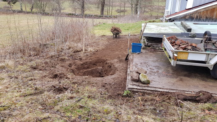 Uppgrävd växtlighet och jord på källarens västsidan med en större sten, skottkärra och släp fullt av tegel och murbruk.