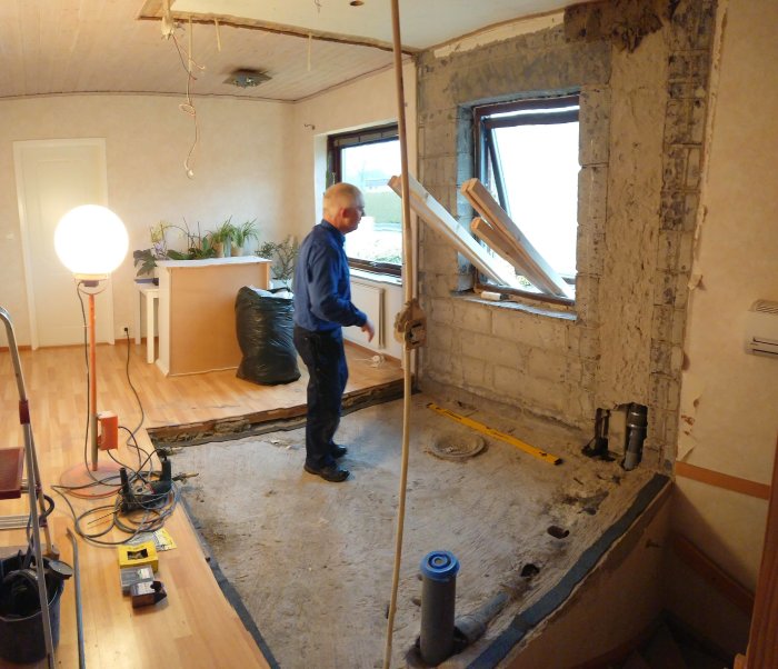 Person i arbetskläder i ett rum under renovering med exponerade betongväggar, verktyg på golvet och en öppning där puts tagits bort.