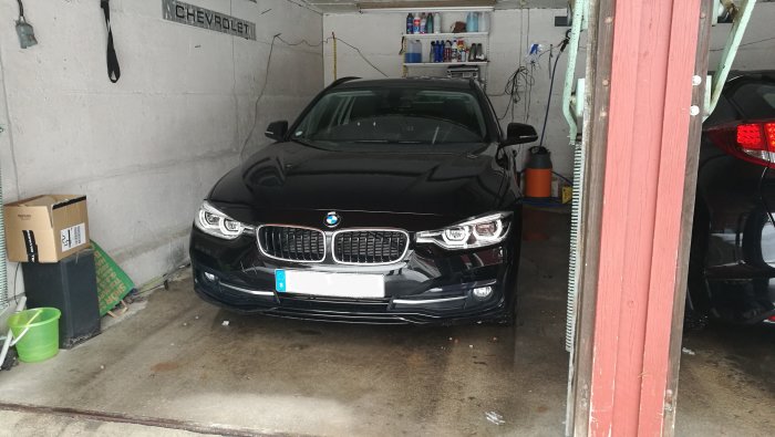 En svart BMW 320d xdrive parkerad i ett garage med diverse föremål och hyllor i bakgrunden.