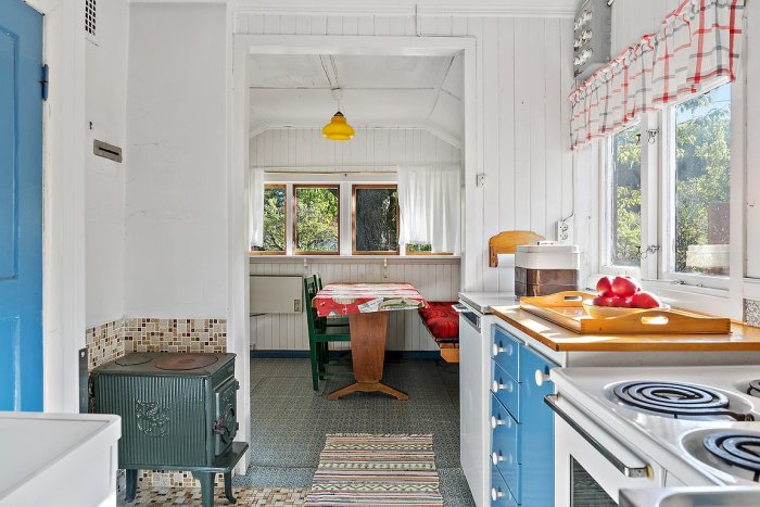 Retrokök i kolonistuga med sprucken Jötul 600 kamin, träbord, inbyggnadsskåp och fönster med röd-vit gardin.
