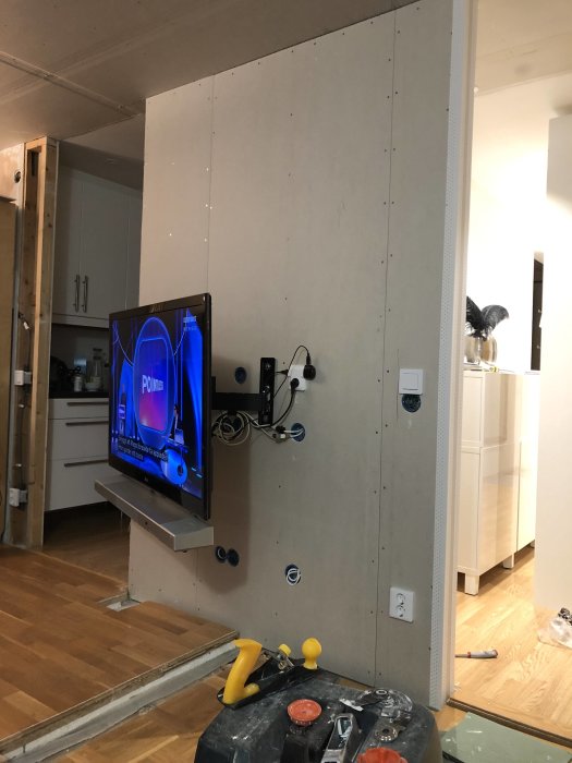 TV monterad på ofärdig vägg med gipsskivor, verktyg och byggmaterial synliga på golvet.