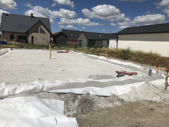 Grunden till en byggnad täckt med stenmjöl, synliga geotextilier och rör, med hus i bakgrunden.