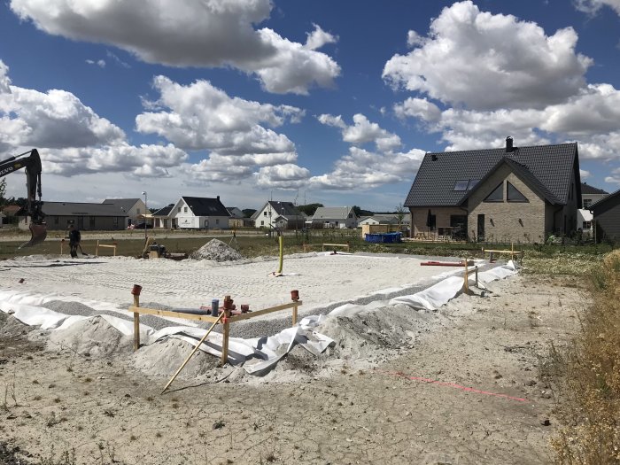 Byggplats med stenmjöl utspritt på marken, hus i bakgrunden och en grävmaskin till vänster.