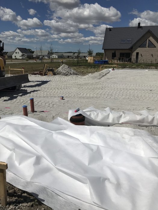 Byggplats med täckt stenmjöl och rör i marken, hus och grävmaskin i bakgrunden.