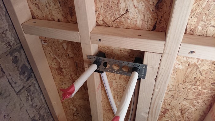 Instalation av rörhållare på en trävägg med osäker fästning enligt instruktioner, med synliga reglar och isolerade rör.