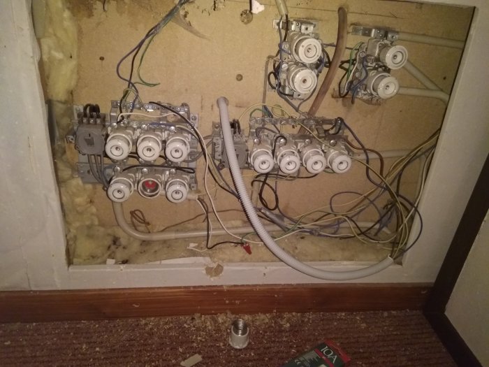 Öppen elcentral med oorganiserade elektriska ledningar och säkringar mot en isolerad vägg.
