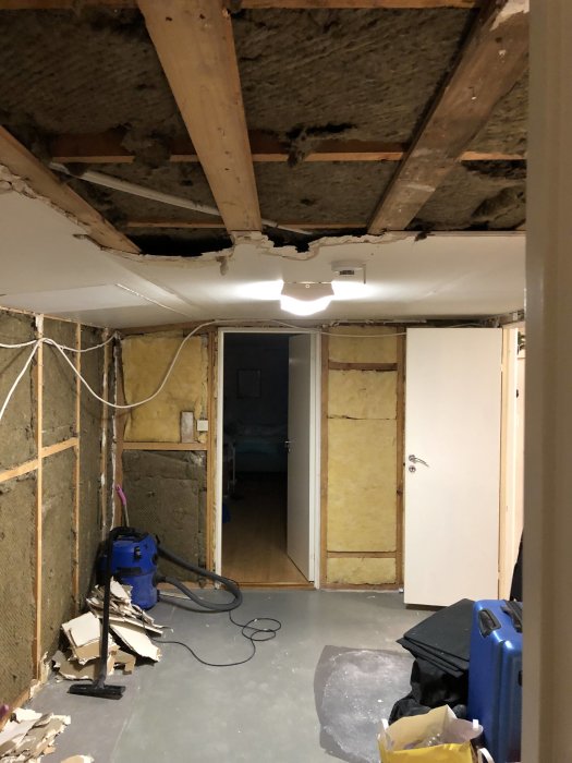 Renoveringsarbete i rum med taket delvis rivet, exponerade träbjälkar och isolering, byggmaterial på golvet.
