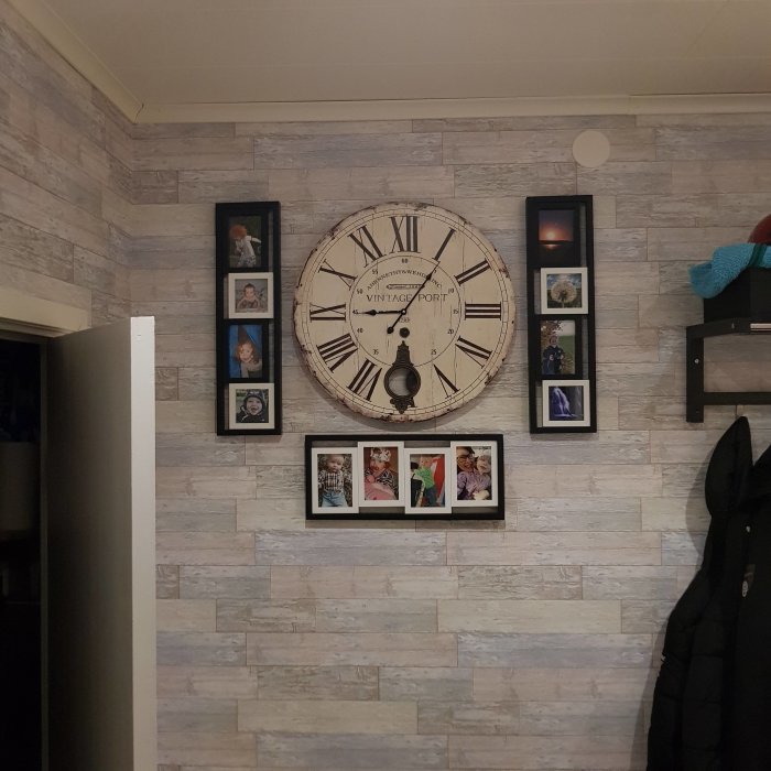 Stor väggklocka omgiven av fotoramar på en grå stenmönstrad vägg i en hall.