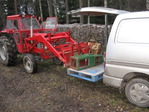 Röd traktor med frontlastare bredvid vit Volvo och en lastpall full med skogsredskap.