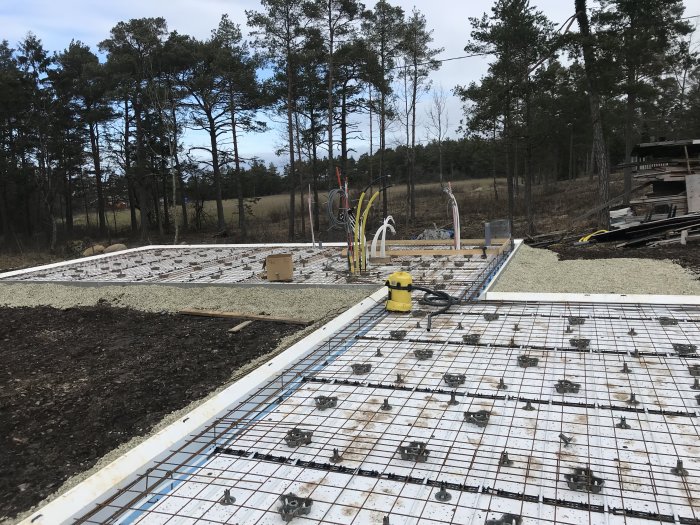 Armerad betonggrund med nät, distanser och rörinstallationer förberedd för gjutning på byggarbetsplats.