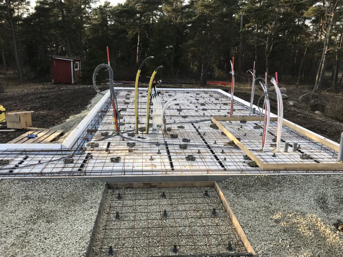 Grundkonstruktion redo för betonggjutning med armeringsnät, slangar och distanser på plats, omgiven av grus och skogsmiljö.