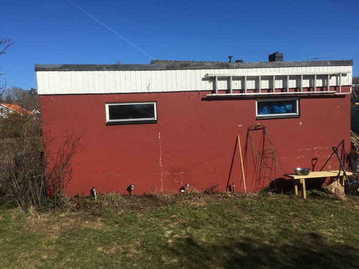 Röd garagevägg med sprickor i putsen, fönster och trädgårdsredskap framför.