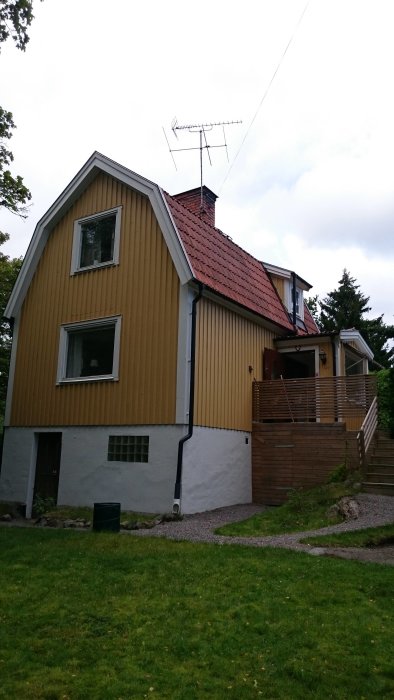 Keltainen puutalon pääty, jossa on punainen tiilikatto ja antenni päällä, kuvattu puutarhasta käsin.