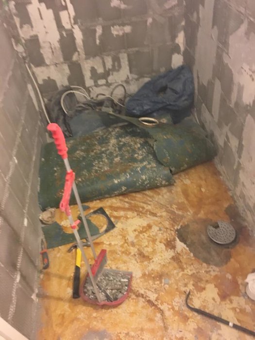 Badrumsgolv med borttagen plastmatta som visar misstänkt fuktskada kring avloppsbrunnen, verktyg och rivningsavfall syns.